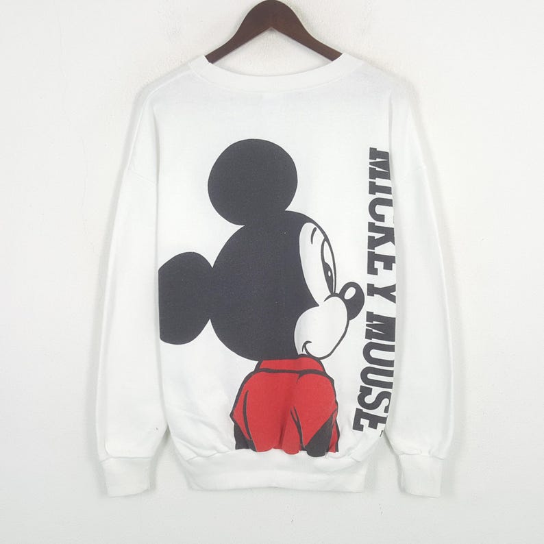 Może przedstawiać: Biała bluza z długim rękawem z dużą grafiką Myszki Miki w kolorach czarnym, czerwonym i białym. Słowa "MICKEY MOUSE" są wydrukowane pionowo na prawym rękawie. Bluza wisi na drewnianym wieszaku.