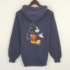 Puede incluir: Sudadera con capucha azul marino con un dibujo de Mickey Mouse en la espalda. El personaje de dibujos animados se muestra en su pose cl&aacute;sica, con pantalones cortos rojos, zapatos amarillos y guantes blancos. La sudadera tiene mangas largas y una capucha.