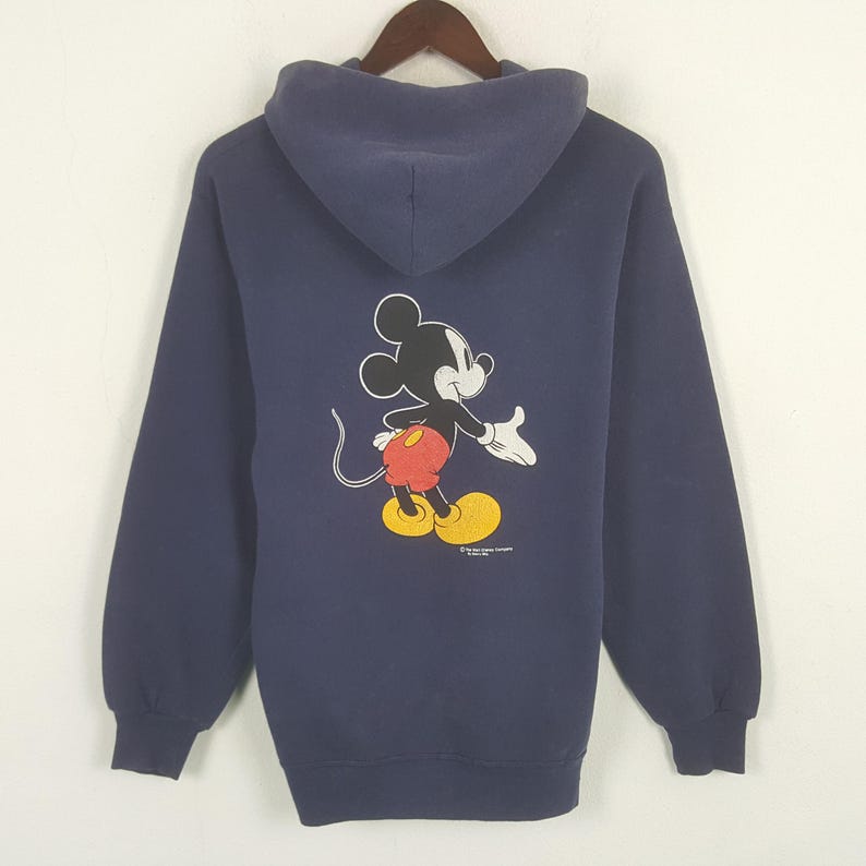 Puede incluir: Sudadera con capucha azul marino con un dibujo de Mickey Mouse en la espalda. El personaje de dibujos animados se muestra en su pose cl&aacute;sica, con pantalones cortos rojos, zapatos amarillos y guantes blancos. La sudadera tiene mangas largas y una capucha.