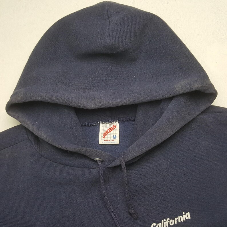 Puede incluir: Sudadera con capucha azul marino con cord&oacute;n y bolsillo delantero. La sudadera tiene una etiqueta que dice "JERZEES" y "M". La palabra "California" est&aacute; impresa en blanco en la parte inferior derecha de la sudadera.