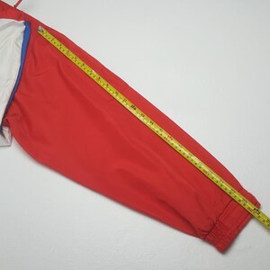 Puede incluir: Chaqueta roja con detalles blancos y azules medida con una cinta m&eacute;trica amarilla. La chaqueta es de material liso y tiene pu&ntilde;o acanalado. La cinta m&eacute;trica muestra una longitud de aproximadamente 152 cm.