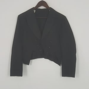 May include: A black formal jacket with a double-breasted front and a unique tailcoat design. The jacket is hanging on a wooden hanger against a plain white background. The jacket has a classic, tailored look.