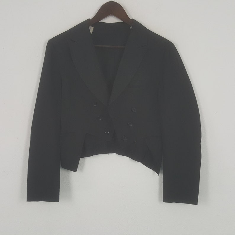 May include: A black formal jacket with a double-breasted front and a unique tailcoat design. The jacket is hanging on a wooden hanger against a plain white background. The jacket has a classic, tailored look.