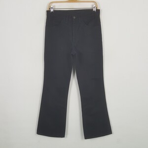 Peut inclure: Pantalon en jean noir avec une coupe évasée. Le pantalon est suspendu à un cintre en bois sur fond blanc. Il possède une fermeture à bouton et fermeture éclair, ainsi que des poches avant et arrière.