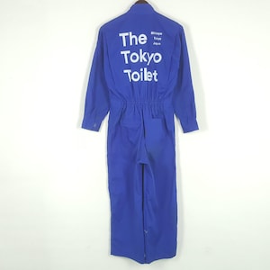 Puede incluir: Mono azul con mangas largas y cierre de botones. La parte trasera del mono presenta el texto "The Tokyo Toilet" en blanco, con "Shibuya Tokyo Japan" impreso encima. El mono tiene cintura fruncida y un bolsillo trasero.