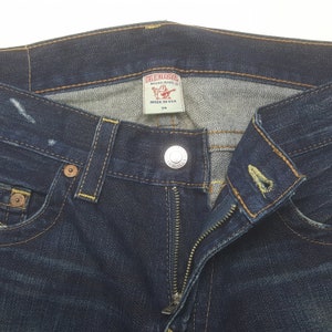 May include: Close-up of dark blue denim jeans with a zip and button fastening. The jeans have a True Religion brand label and a size 34 tag. Yellow stitching and metal button and rivets are visible.