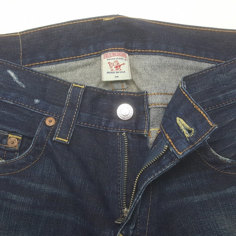 May include: Close-up of dark blue denim jeans with a zip and button fastening. The jeans have a True Religion brand label and a size 34 tag. Yellow stitching and metal button and rivets are visible.
