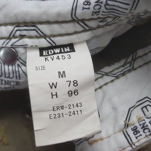 May include: Close-up of a white tag with black text from the brand Edwin. The tag displays the size M, with measurements W 78 and H 96. The tag is attached to a pair of jeans. The jeans are olive green.