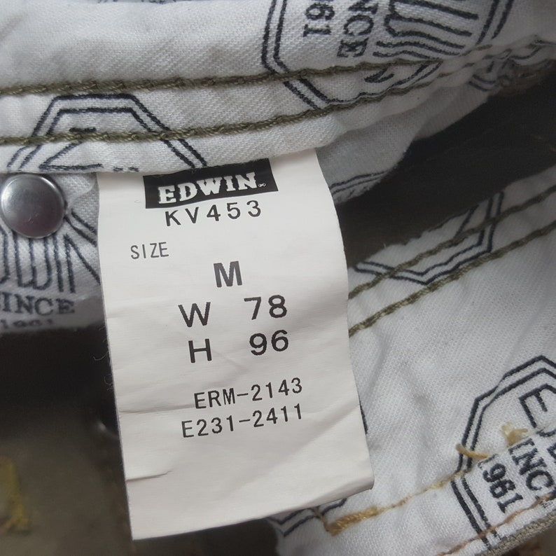 May include: Close-up of a white tag with black text from the brand Edwin. The tag displays the size M, with measurements W 78 and H 96. The tag is attached to a pair of jeans. The jeans are olive green.