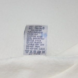 May include: A white fabric tag with black text detailing garment care instructions. The tag includes information on washing, drying, and ironing, along with the fabric composition: 50% cotton, 50% Trevira polyester. The tag also has the RN number TI908.