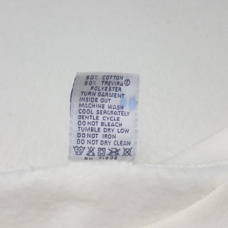 May include: A white fabric tag with black text detailing garment care instructions. The tag includes information on washing, drying, and ironing, along with the fabric composition: 50% cotton, 50% Trevira polyester. The tag also has the RN number TI908.