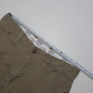 May include: Khaki-colored pants with a button closure and a measuring tape laid across the top. The pants have visible stitching and a patterned interior lining. The measuring tape shows a length of approximately 18 inches.