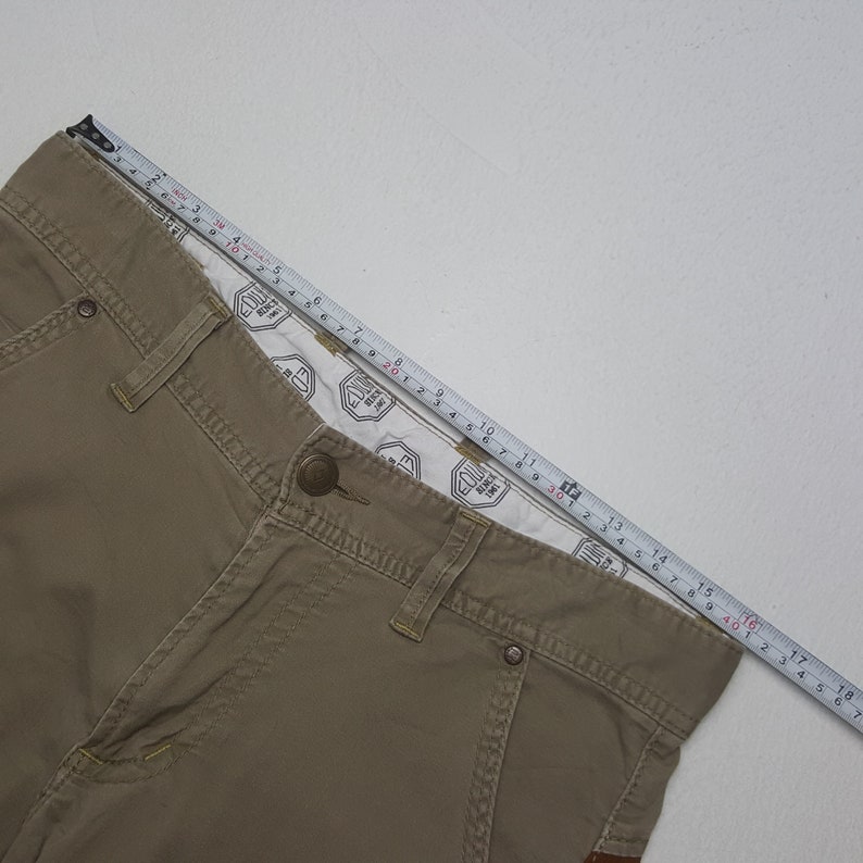 May include: Khaki-colored pants with a button closure and a measuring tape laid across the top. The pants have visible stitching and a patterned interior lining. The measuring tape shows a length of approximately 18 inches.