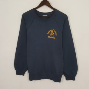Puede incluir: Sudadera azul marino con el texto "Raymond Central Mustangs" en amarillo. La sudadera tiene mangas largas y cuello redondo. La prenda está colgada de una percha de madera.