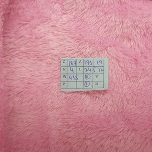 May include: Close-up of a soft, fluffy pink fabric. A small white tag with handwritten numbers and letters is visible in the upper right corner. The fabric has a dense, textured appearance.