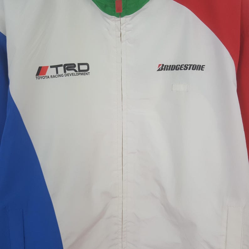 Puede incluir: Chaqueta de carreras blanca, azul y roja con los logotipos "TRD TOYOTA RACING DEVELOPMENT" y "BRIDGESTONE". La chaqueta tiene una cremallera completa y un cuello alto. Las mangas son azules y rojas.