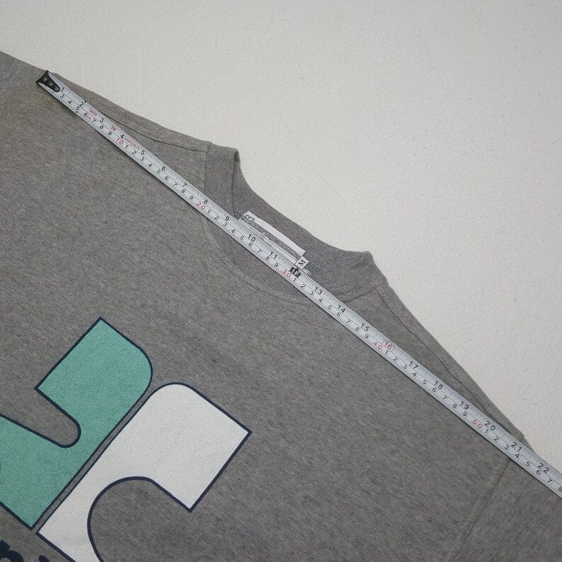 Pode incluir: Uma camisola cinza de gola redonda com um grande design gr&aacute;fico turquesa e branco. Uma fita m&eacute;trica est&aacute; colocada sobre a camisola, mostrando medidas em cent&iacute;metros. A camisola est&aacute; sobre uma superf&iacute;cie branca.