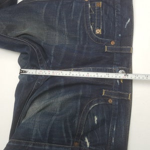May include: Dark blue denim jeans with a faded wash and distressed details. The jeans feature classic five-pocket styling, yellow stitching, and metal rivets. A measuring tape shows a waist measurement of approximately 30 cm.