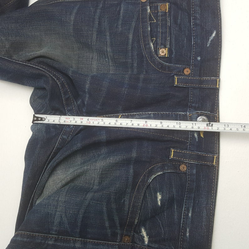 May include: Dark blue denim jeans with a faded wash and distressed details. The jeans feature classic five-pocket styling, yellow stitching, and metal rivets. A measuring tape shows a waist measurement of approximately 30 cm.