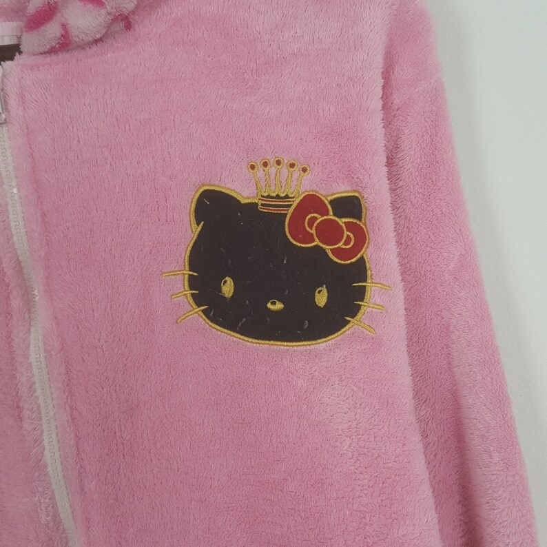 May include: A pink, plush zip-up hoodie featuring a Hello Kitty graphic. The graphic includes a brown Hello Kitty face with a red bow and gold crown. The hoodie has a white zipper and a fluffy collar.