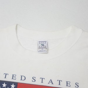 May include: A white crew neck sweatshirt with the text "UNITED STATES" in navy blue. The sweatshirt features a red and blue graphic at the bottom. The tag reads "GALT SAND" and "MADE IN U.S.A."