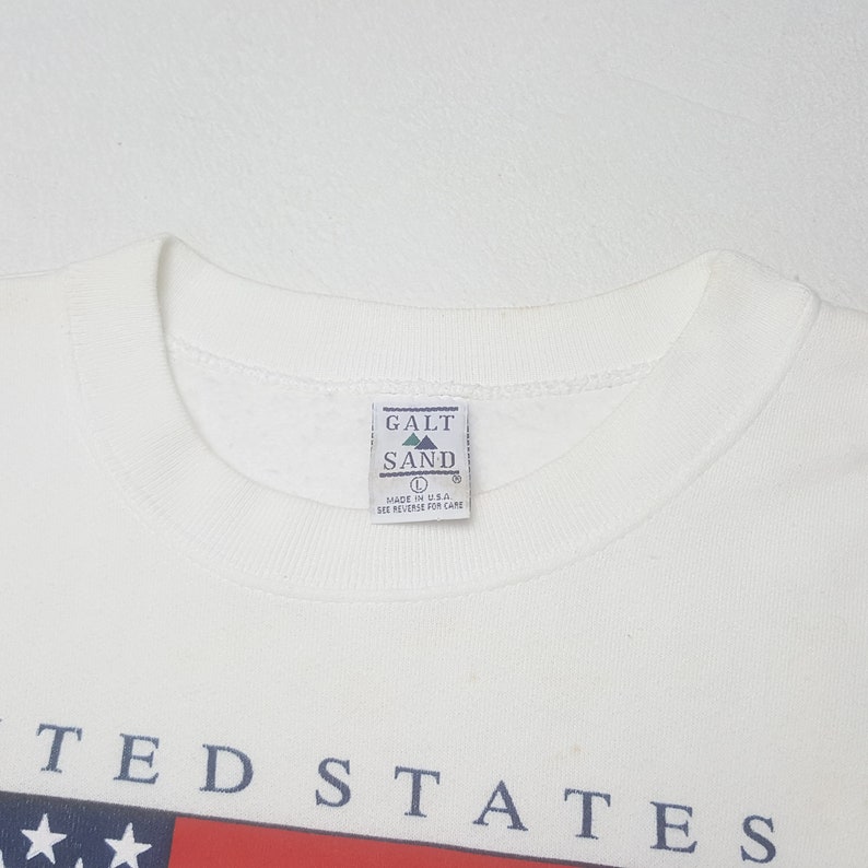 May include: A white crew neck sweatshirt with the text "UNITED STATES" in navy blue. The sweatshirt features a red and blue graphic at the bottom. The tag reads "GALT SAND" and "MADE IN U.S.A."