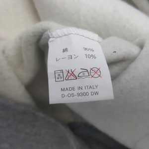Pode incluir: Close-up de uma etiqueta de roupa com caracteres japoneses e a inscri&ccedil;&atilde;o "MADE IN ITALY". A etiqueta indica uma composi&ccedil;&atilde;o de tecido de 90% algod&atilde;o e 10% rayon. As instru&ccedil;&otilde;es de lavagem tamb&eacute;m s&atilde;o vis&iacute;veis. A etiqueta est&aacute; presa a uma pe&ccedil;a de roupa cinza e branca.
