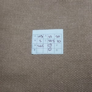 Puede incluir: Primer plano de una tela beige con una etiqueta blanca. La etiqueta tiene texto y n&uacute;meros en negro, incluyendo "C 103", "A 22" y "L 28.5". La tela tiene una textura tejida.