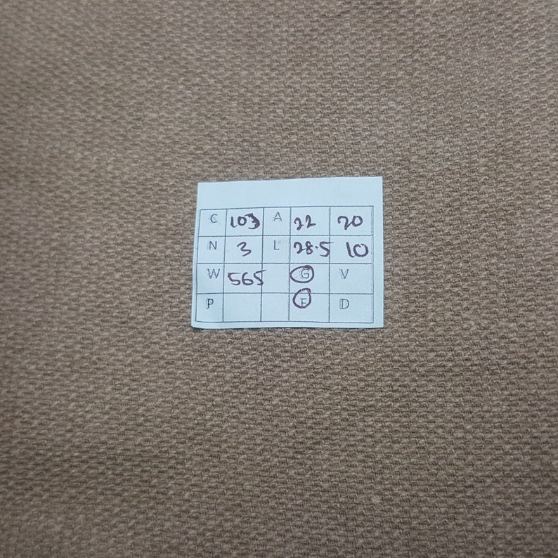 Puede incluir: Primer plano de una tela beige con una etiqueta blanca. La etiqueta tiene texto y n&uacute;meros en negro, incluyendo "C 103", "A 22" y "L 28.5". La tela tiene una textura tejida.