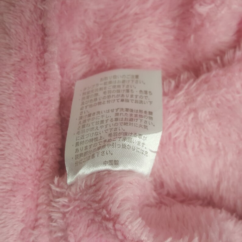 May include: Close-up of a pink, fluffy fabric with a white tag attached. The tag has Japanese text and is likely a care label. The fabric appears soft and plush, suggesting a garment or textile product.