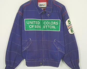 May include: A vintage blue jacket with a grid pattern and the text "UNITED COLORS OF BEN ETTON." The jacket has a collar, pockets, and a patch with the numbers "5" and "6." Red trim accents the shoulders. The jacket is hanging on a wooden hanger.