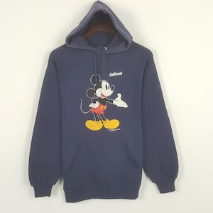 Puede incluir: Sudadera con capucha azul marino con un dibujo de Mickey Mouse. Mickey lleva pantalones cortos rojos y zapatos amarillos. La palabra "California" est&aacute; impresa encima del dibujo. La sudadera tiene un bolsillo delantero.