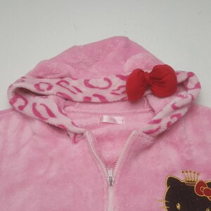 May include: Pink hooded onesie with a zipper closure. The hood features a red bow and a pink and white leopard print design. The onesie has a Hello Kitty graphic on the front.