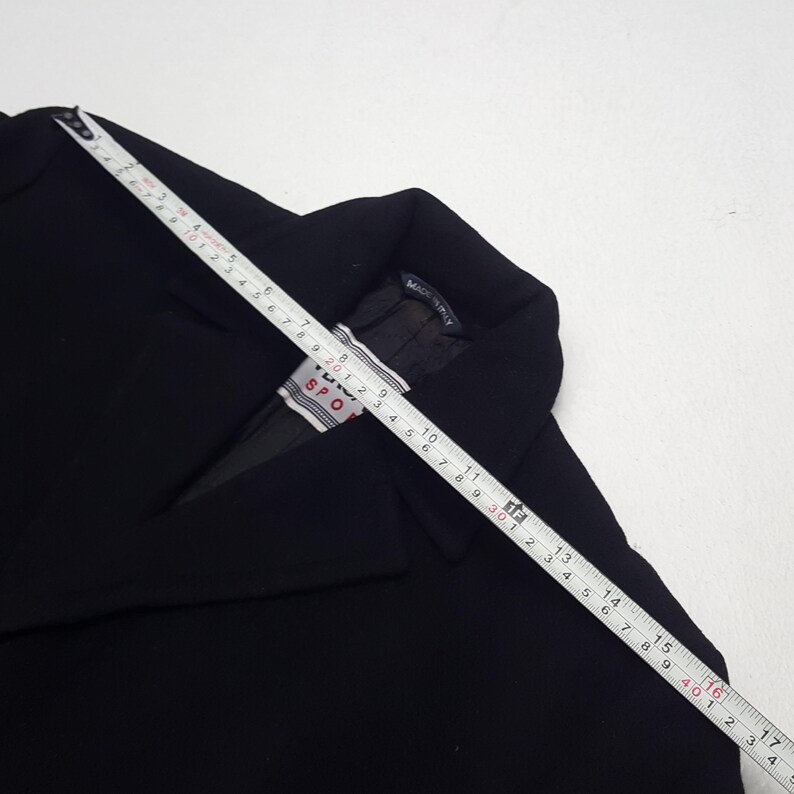 Może przedstawiać: Czarny blazer z miarką na wierzchu. Blazer ma metkę z napisem "TUV SPORT". Miarka pokazuje pomiary w centymetrach. Blazer jest wyprodukowany we Włoszech.