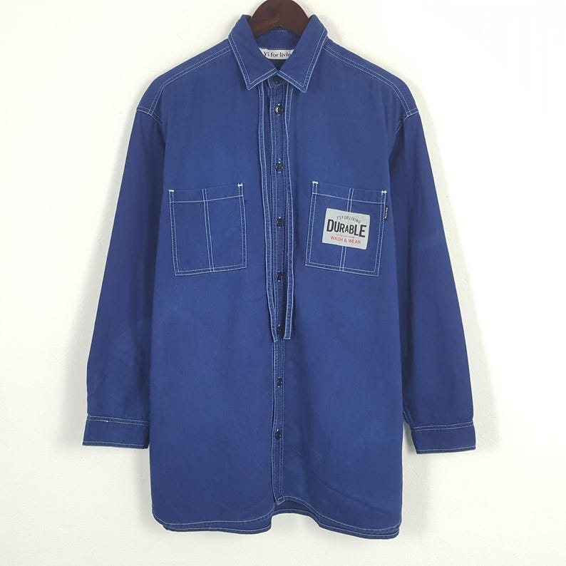 May include: A long-sleeved, cobalt blue shirt with white stitching details. Features two front pockets and a rectangular patch that reads "Durable Wash & Wear". The shirt is button-down with a collared neckline.