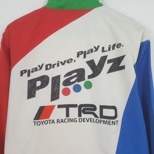 Puede incluir: Chaqueta blanca, roja y azul con el texto "Play Drive, Play Life" y "Playz" en negrita. La chaqueta tambi&eacute;n presenta el logotipo "TRD TOYOTA RACING DEVELOPMENT". La chaqueta tiene un cuello verde.
