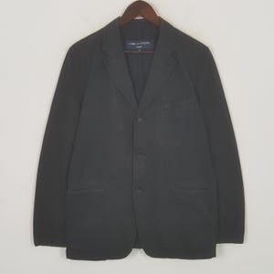 Puede incluir: Un blazer negro con tres botones y dos bolsillos delanteros, colgado de una percha de madera. El blazer tiene solapas con muescas y mangas largas. La marca "COMME des GAR&Ccedil;ONS HOMME" es visible en la etiqueta.