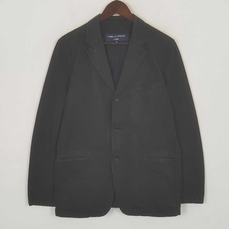 Puede incluir: Un blazer negro con tres botones y dos bolsillos delanteros, colgado de una percha de madera. El blazer tiene solapas con muescas y mangas largas. La marca "COMME des GAR&Ccedil;ONS HOMME" es visible en la etiqueta.