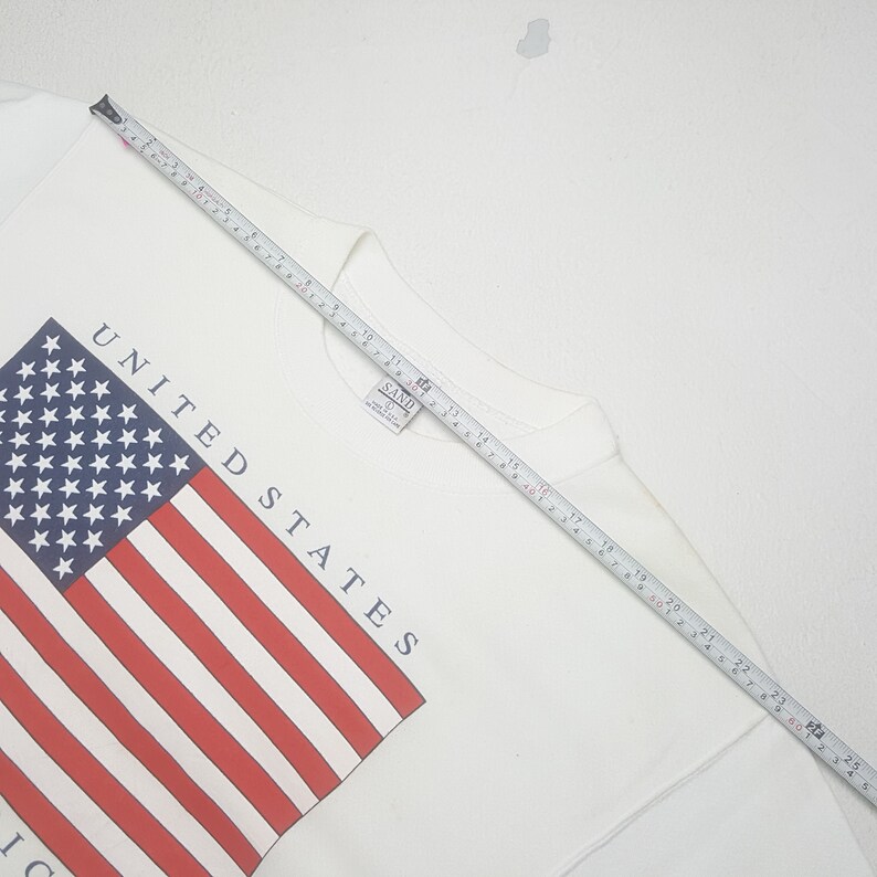 May include: A white sweatshirt featuring the American flag design. The flag is red, white, and blue, with the words "UNITED STATES" printed in navy blue. The sweatshirt is measured with a tape measure.