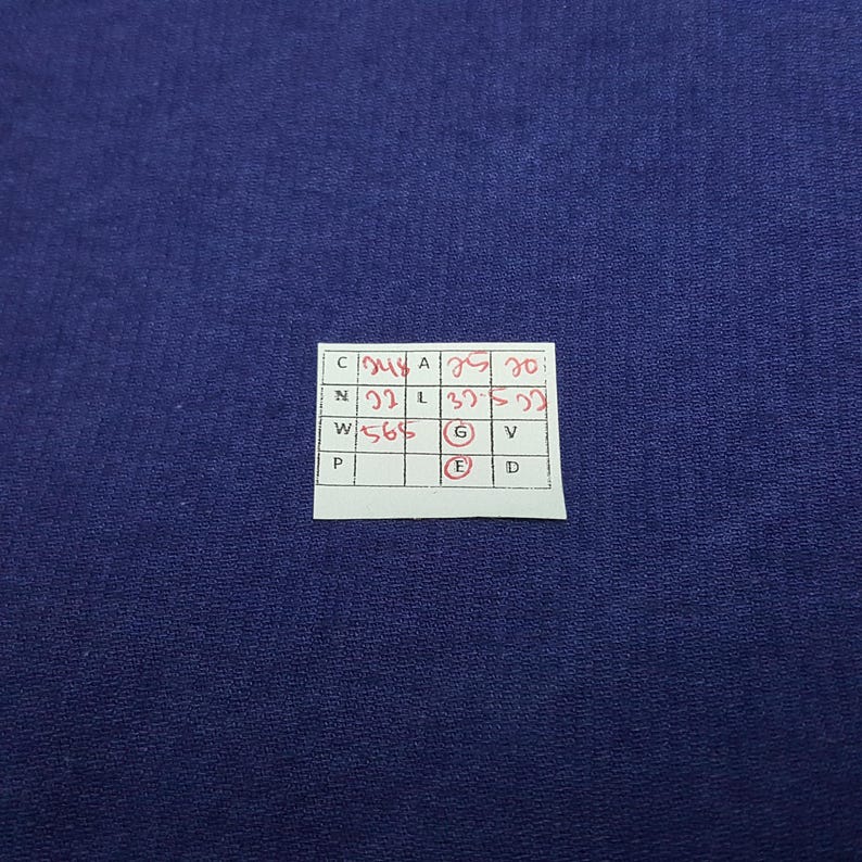 May include: A close-up shot of a dark blue fabric. A small white tag with handwritten red text is in the lower right corner. The tag has a grid of boxes with letters and numbers.