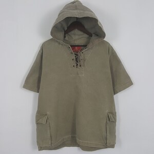 Puede incluir: Camiseta con capucha verde oliva con mangas cortas. La camiseta presenta un escote con cordones y dos bolsillos delanteros. La capucha est&aacute; adjunta y la camiseta est&aacute; colgada en una percha de madera.