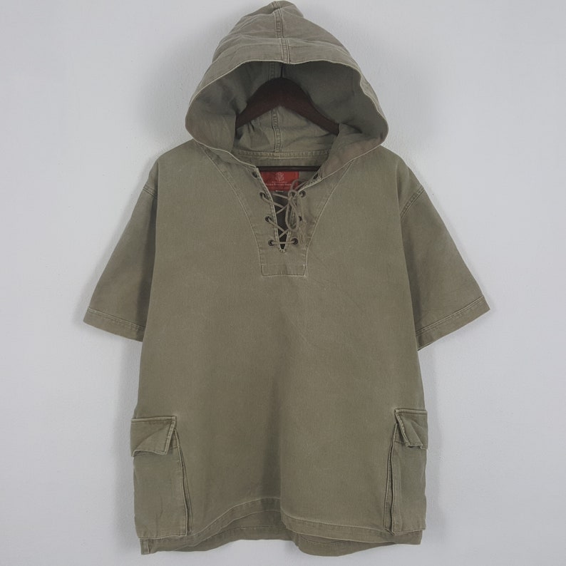 Puede incluir: Camiseta con capucha verde oliva con mangas cortas. La camiseta presenta un escote con cordones y dos bolsillos delanteros. La capucha est&aacute; adjunta y la camiseta est&aacute; colgada en una percha de madera.