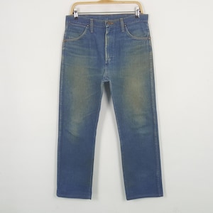 May include: Blue denim jeans on a wooden hanger. The jeans feature a classic five-pocket design, button and zip closure, and a faded wash. The straight-legged jeans appear to be vintage, with a worn look.