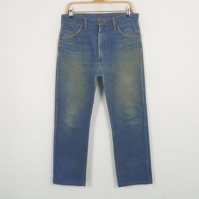 May include: Blue denim jeans on a wooden hanger. The jeans feature a classic five-pocket design, button and zip closure, and a faded wash. The straight-legged jeans appear to be vintage, with a worn look.
