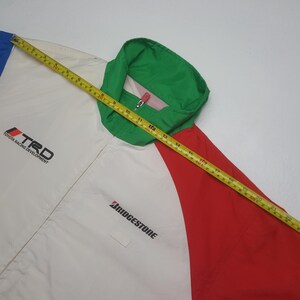 Puede incluir: Chaqueta multicolor con cuerpo blanco, detalles en rojo y azul y cuello verde. La chaqueta presenta los logotipos "TRD Toyota Racing Development" y "Bridgestone". Una cinta m&eacute;trica amarilla est&aacute; colocada sobre la chaqueta.