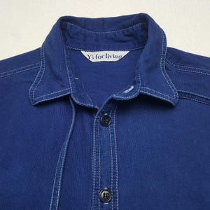 May include: Close-up of a navy blue button-down shirt with white stitching details. The shirt features a collar and two black buttons. The label reads "Y's for living".