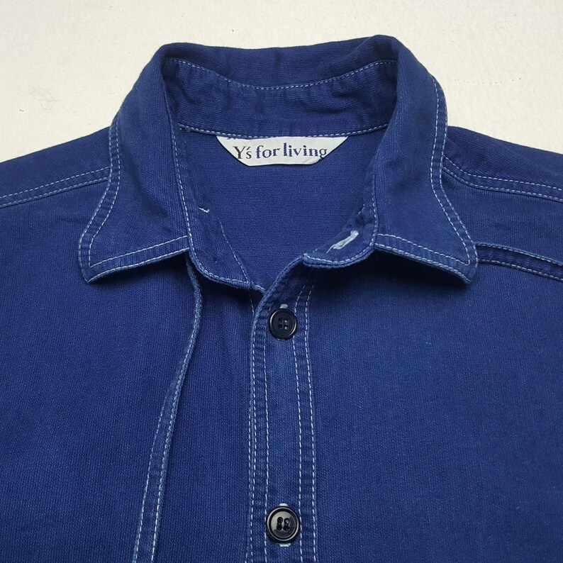 May include: Close-up of a navy blue button-down shirt with white stitching details. The shirt features a collar and two black buttons. The label reads "Y's for living".