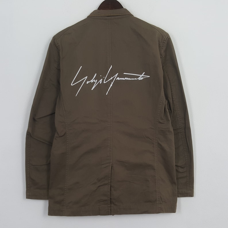 Puede incluir: Blazer verde oliva con un logotipo de firma blanco en la espalda. La chaqueta tiene un cuello cl&aacute;sico y cierre de botones. La chaqueta de manga larga est&aacute; colgada en una percha de madera.