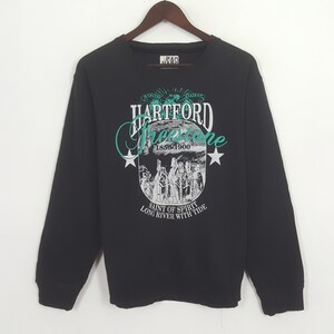 May include: Black sweatshirt with a graphic design featuring the text "Hartford Freestone" in white and teal, along with an illustration and the text "Saint of Spirit Long River With Tide". The sweatshirt is hanging on a wooden hanger.