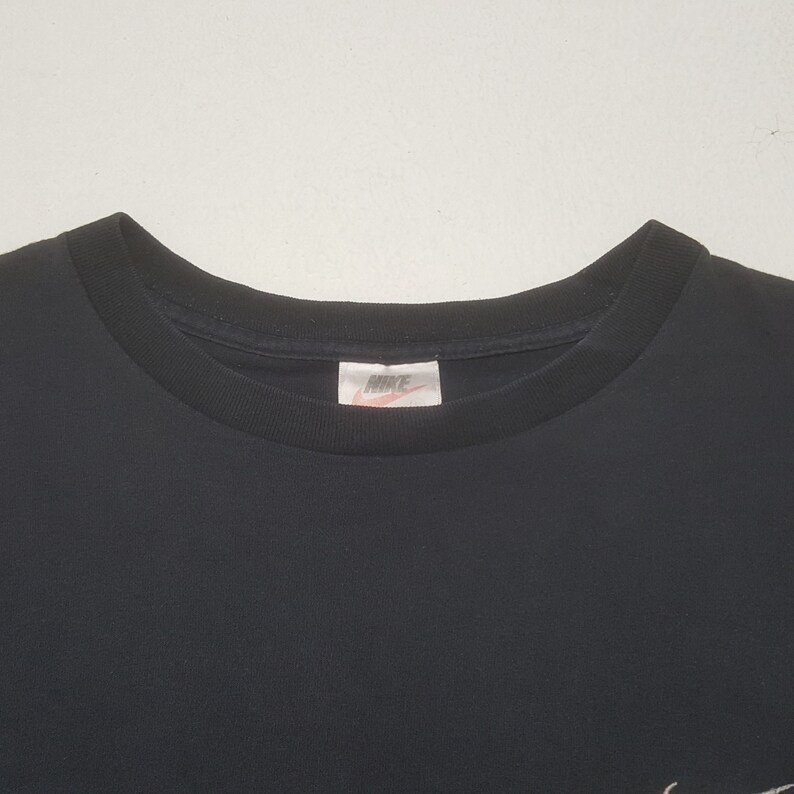 Peut inclure: T-shirt noir &agrave; col rond avec le logo Nike sur l'&eacute;tiquette. Le t-shirt est uni et a un design simple. Le logo Nike est blanc avec un swoosh rouge.